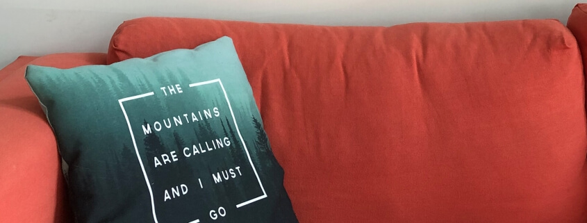 Wide image with a red couch and a pillow which says "The mountains are calling I must go"